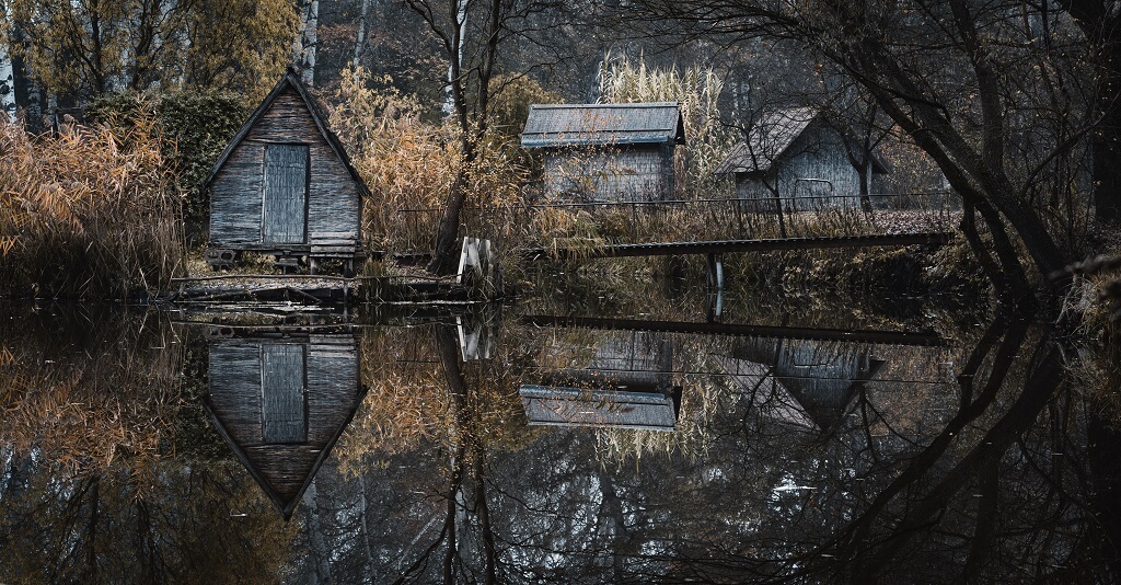 A dark, dreary scene of three wooden sheds and a bridge in a dark woods reflecting in a pond, representing how self criticism darkens our self reflection.