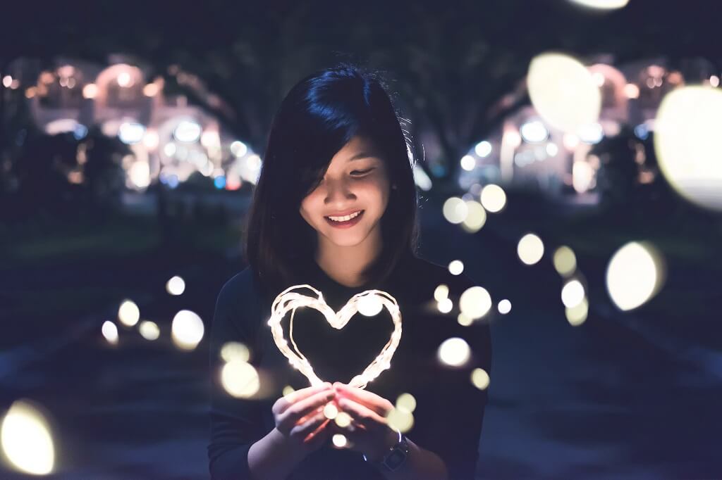 Consider the content of your heart as you see a young woman smiling as she looks down at glowing lights in the shape of a heart. 