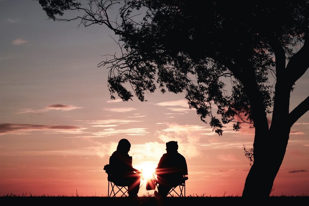 The photo shows silhouette of two friends sitting near a tree at sunset. 