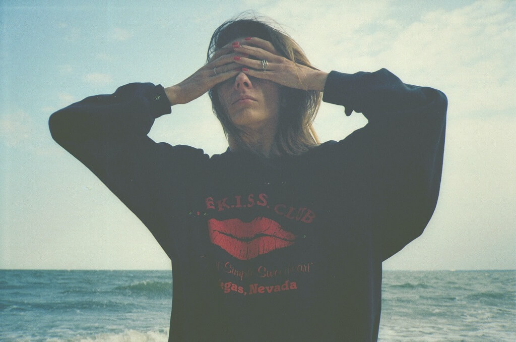 The photo shows a young woman standing with her back to the ocean, covering her eyes with her hands. How will she say something if she tries not to see.