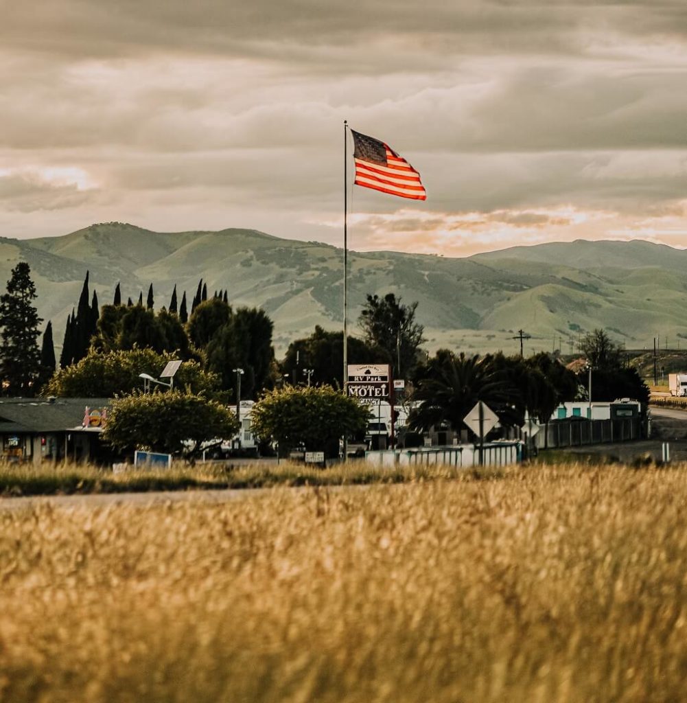 Photo shows an American flag waving above beautiful farmland and small town as we reflect on how to honor our heroes.