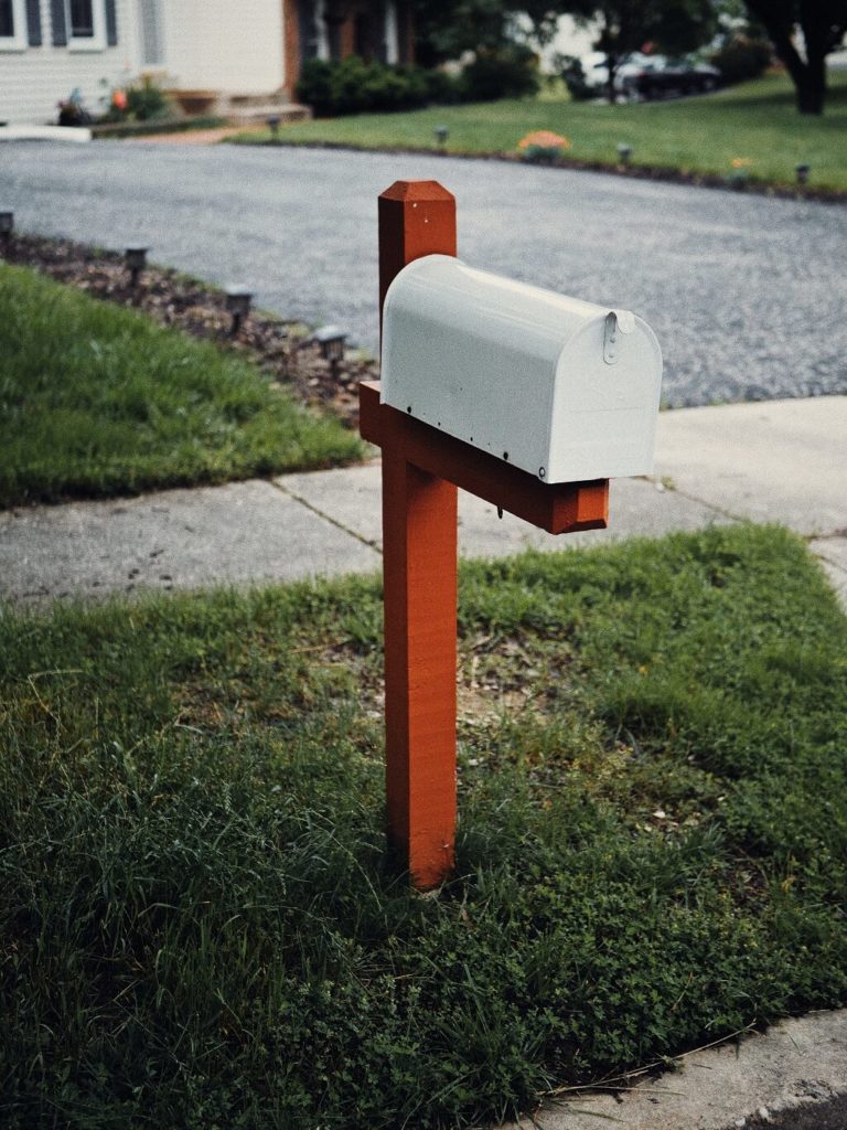 Photo shows a grey metal mail box on a wooden stand,  set in the grass near a sidewalk by the side of a neighborhood street. Imagine your Valentine in the mail box.
